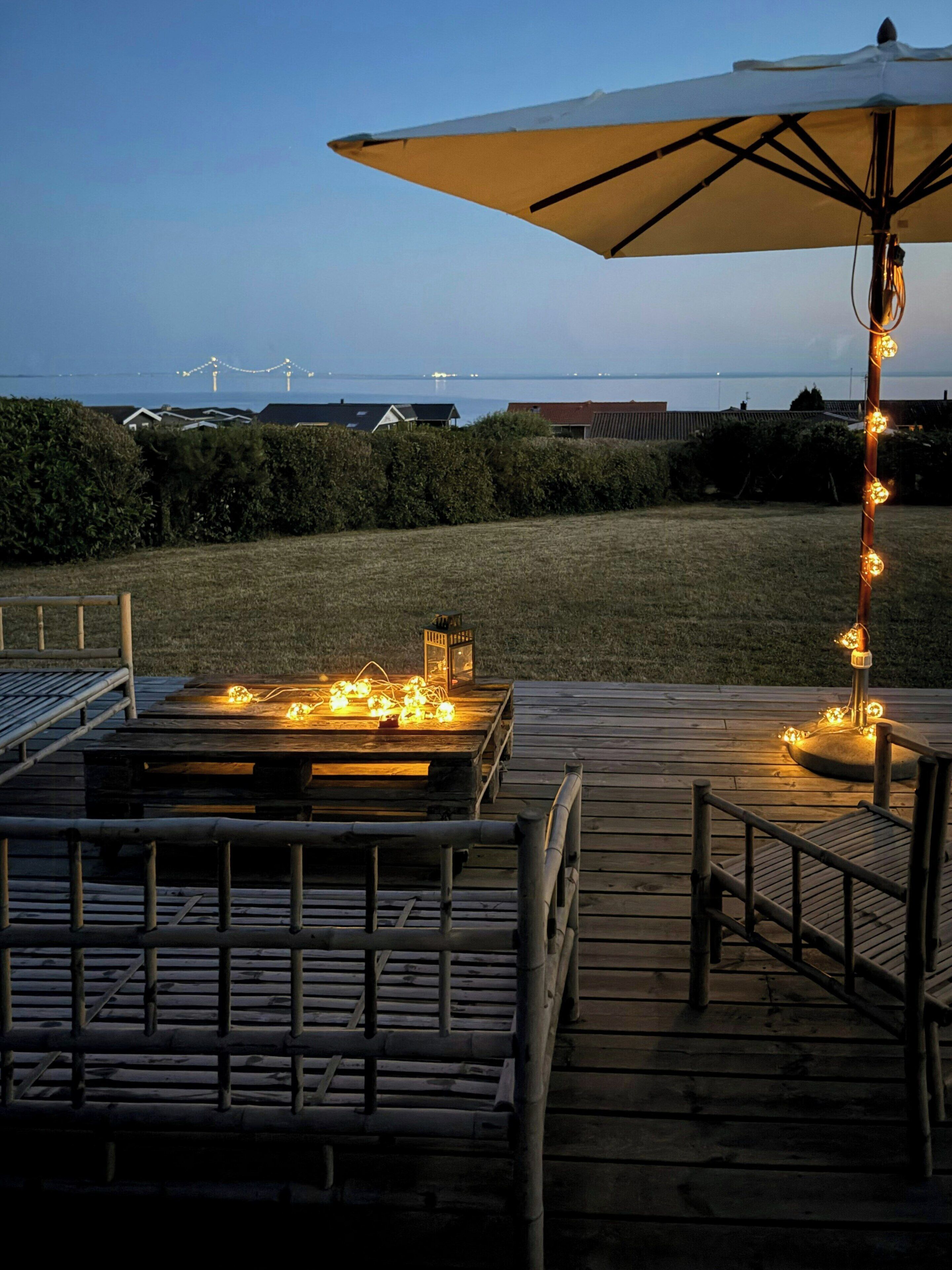 An outdoor deck with a table and chairs, a string of lights, and a view of the sea and wind turbines in the background.