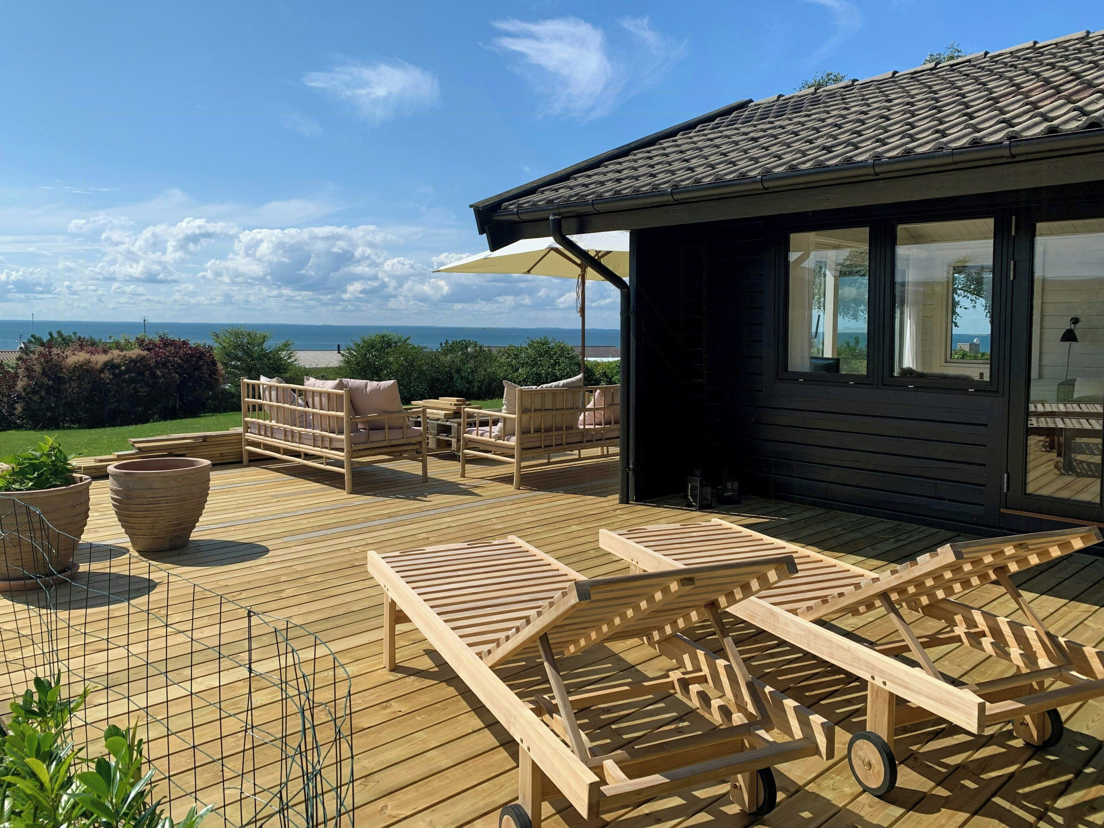 A wooden deck with outdoor furniture, a view of the sea, and a building with large windows.