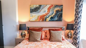 Ein Bett mit floralem Überwurf, zwei Zierkissen, ein an der Wand hängendes abstraktes Gemälde und zwei Nachttischlampen.