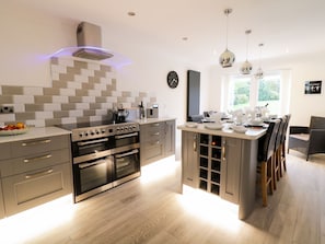 A modern kitchen with a central island, built-in ovens, and a large window providing natural light. - Gwelfor (Cemaes Bay)