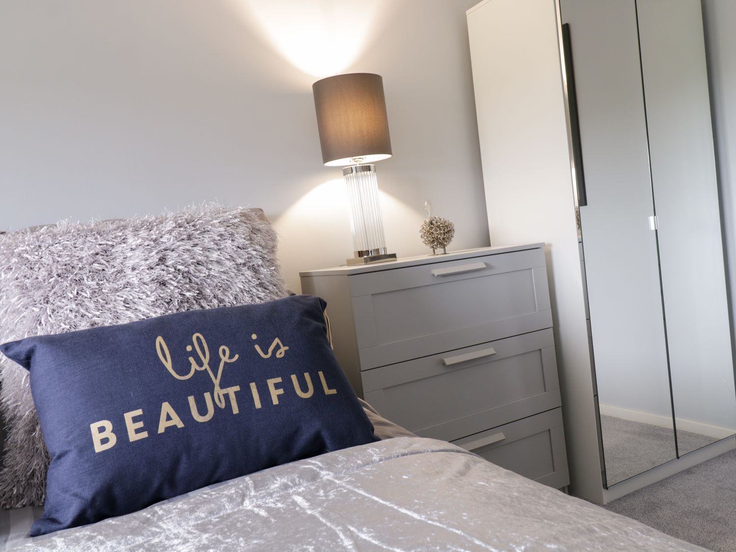 Un dormitorio con cama, mesita de noche, lámpara, armario y un cojín decorativo.