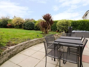 A patio with a table and chairs, surrounded by a brick wall and a grassy area with various plants. - Gwelfor (Cemaes Bay)