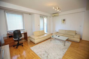 Un salon moderne avec un plancher en bois, un canapé beige, un fauteuil de bureau noir, une petite table basse et une grande fenêtre qui laisse entrer la lumière naturelle.