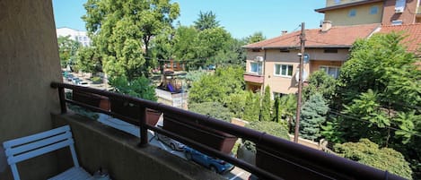 A balcony with a white bench and a table, overlooking a street with parked cars and buildings.