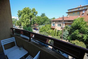 Un balcon avec un banc blanc et une table, donnant sur une rue bordée de voitures garées et d’immeubles.