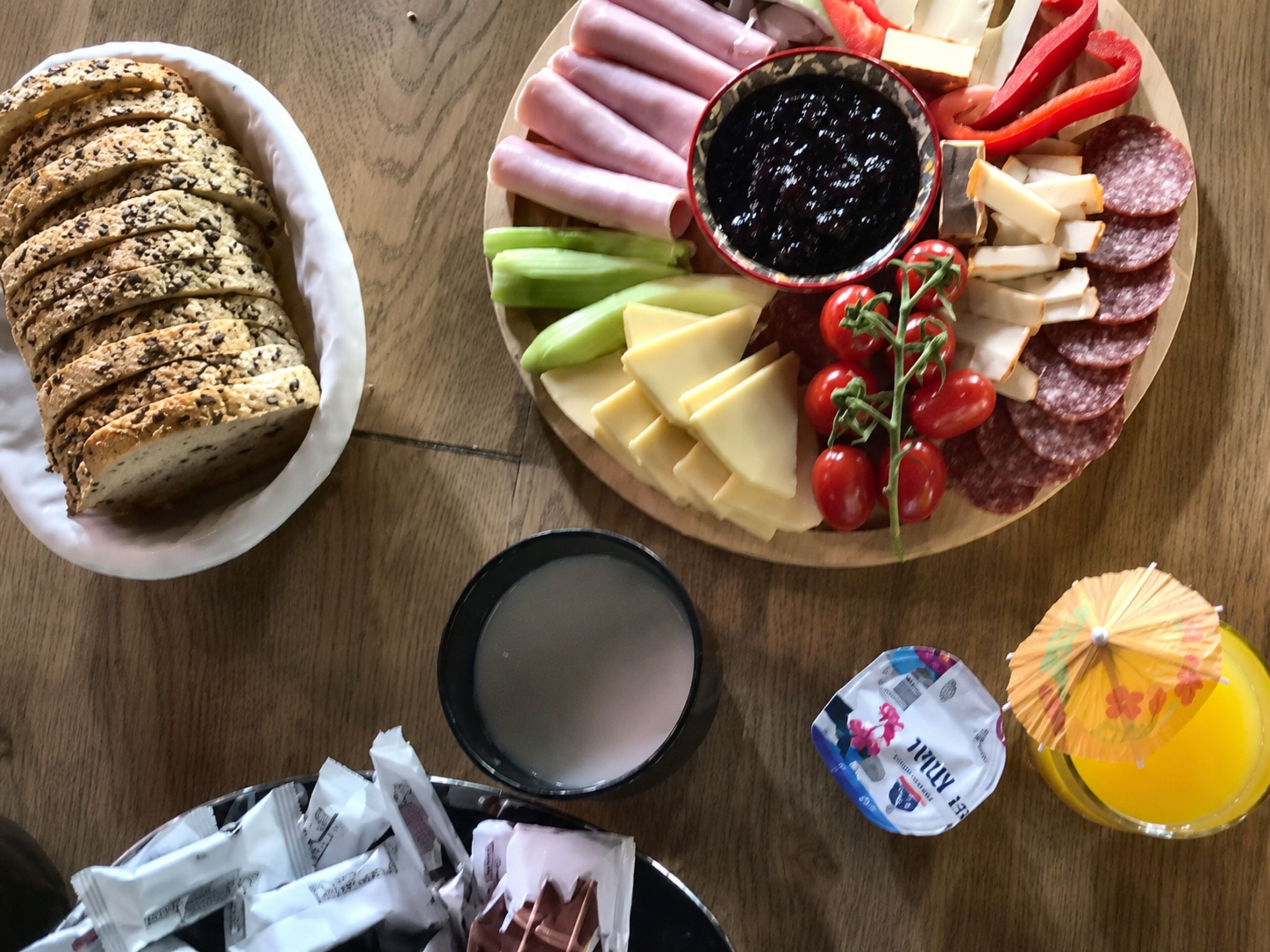 A wooden table with a variety of food items including sliced bread, cheese, meats, and a bowl of jam.