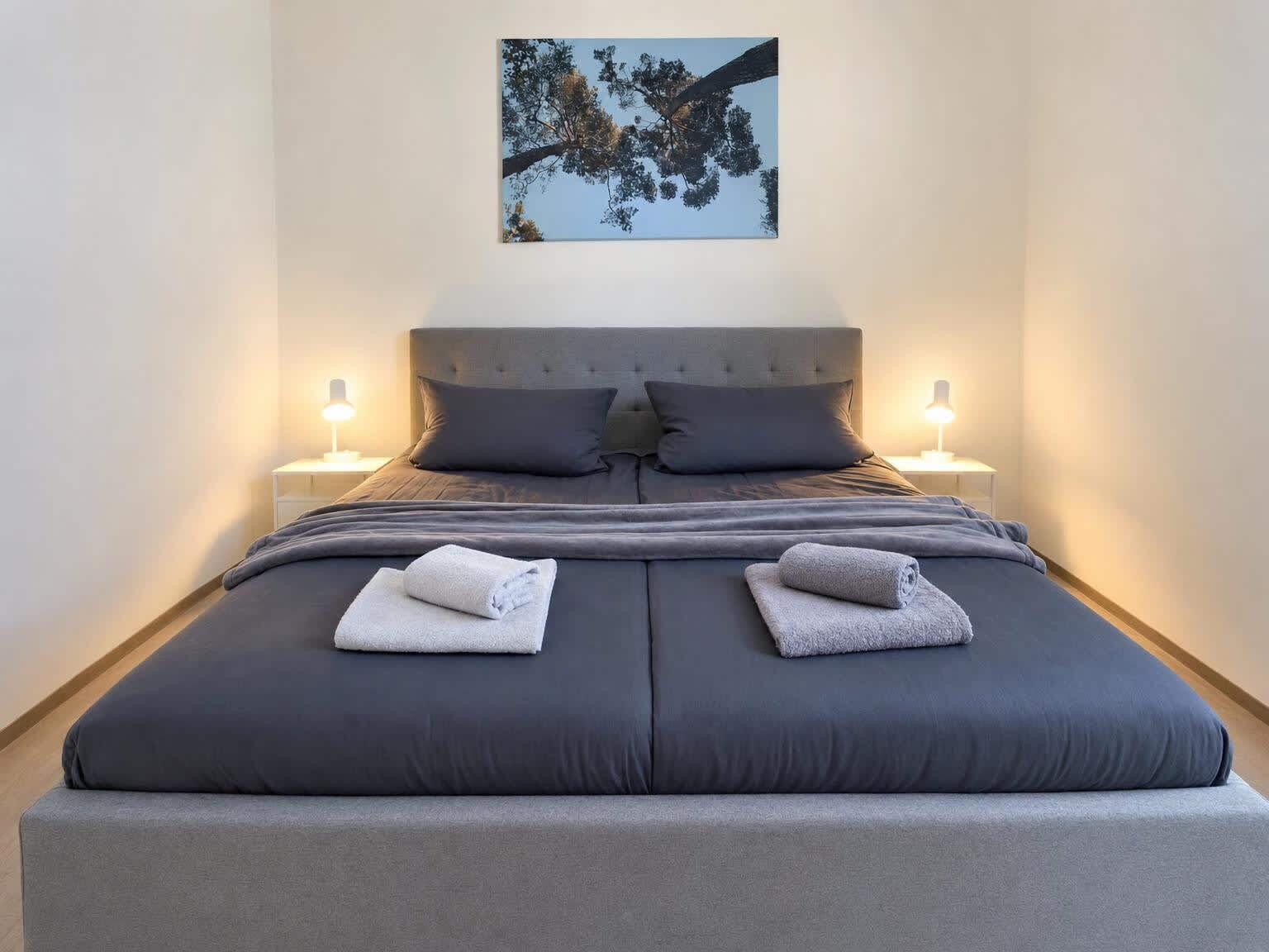 Een net opgemaakt bed met twee kussens, twee handdoeken gevouwen en twee bedlampjes.