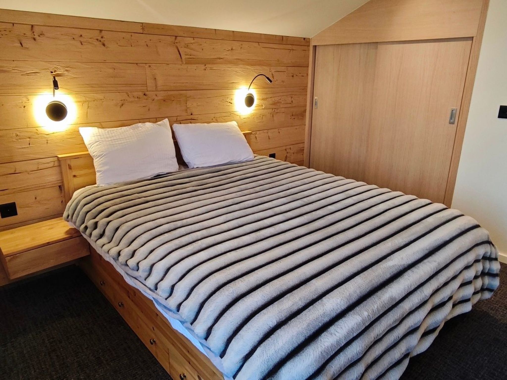 A bed with a striped blanket, wooden headboard, two bedside lamps, and a wooden wardrobe.
