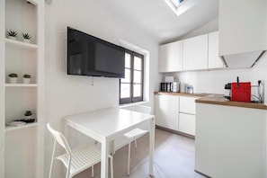 A compact kitchen with white cabinetry, a flat-screen TV mounted above a window, a small dining table with a chair, and a red appliance on the counter. - A/C studio in the heart of Cannes – Old Port (Cannes)
