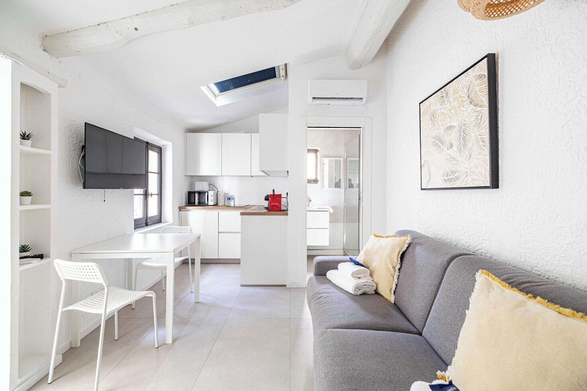 Eine moderne Küche mit weißen Schränken, ein Essbereich mit weißem Tisch und Stühlen, ein graues Sofa mit Kissen und ein gerahmtes Kunstwerk an der Wand.