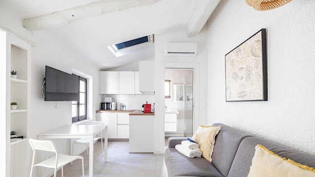 Eine moderne Küche mit weißen Schränken, ein Essbereich mit weißem Tisch und Stühlen, ein graues Sofa mit Kissen und ein gerahmtes Kunstwerk an der Wand.