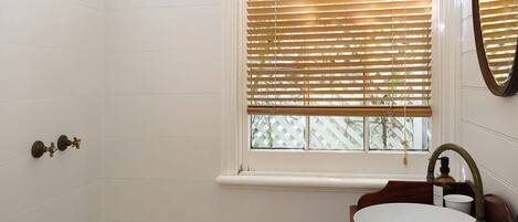 A bathroom with a shower, a sink with a wooden vanity, a window with blinds, and a mirror.