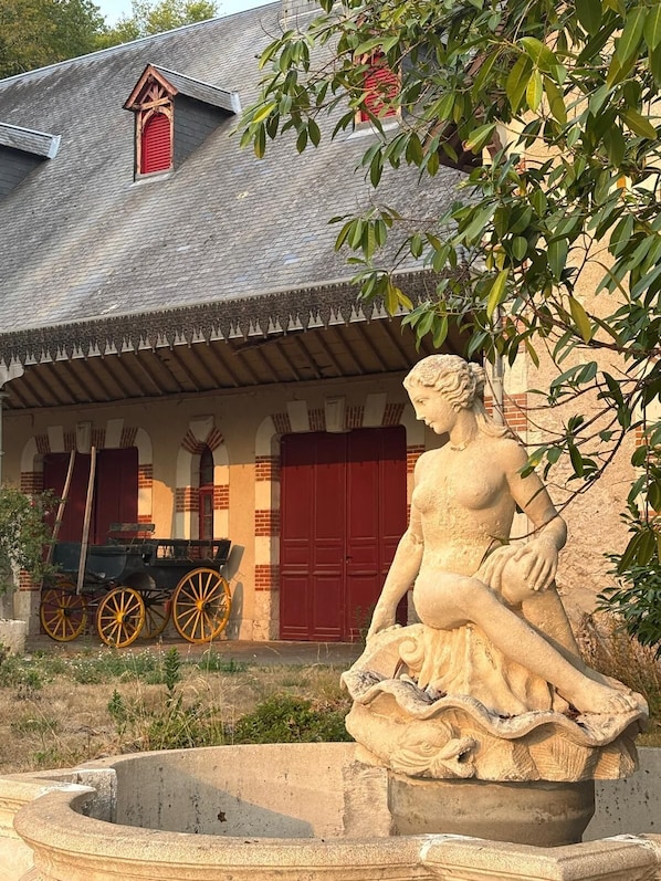 A stone statue of a woman seated on a pedestal, with a building featuring a red door and a cart in the background.