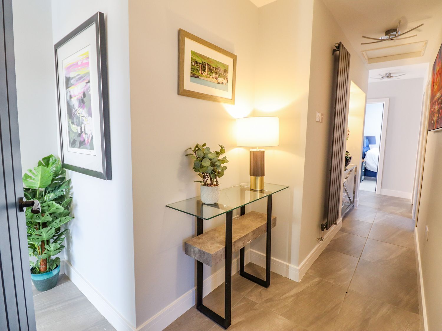 A hallway with a glass console table, a potted plant, a framed picture, and a lamp.