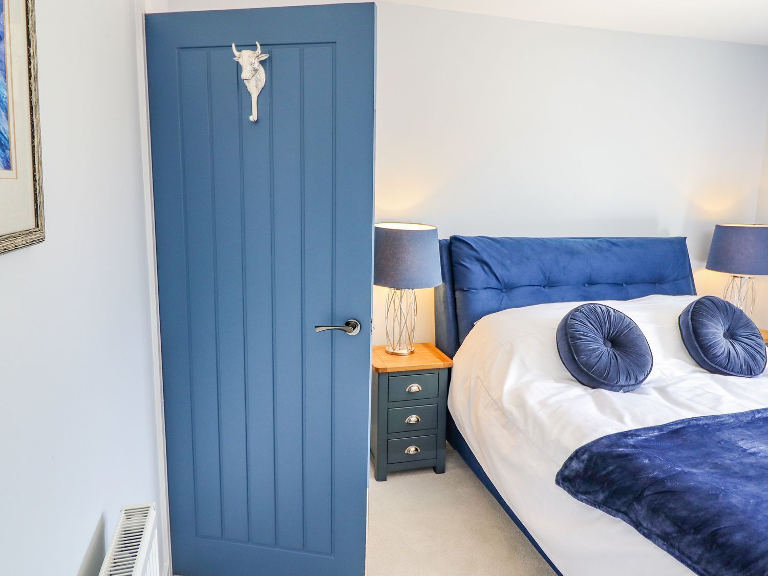 A bedroom with a blue door, a bed with white bedding and two blue pillows, a bedside table with a lamp, and a small drawer unit.