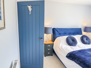 A bedroom with a blue door, a bed with white bedding and two blue pillows, a bedside table with a lamp, and a small drawer unit. - Trefin (Cemaes Bay)
