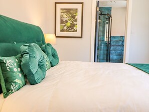 Una cama con almohadas verdes y un edredón blanco, un cuadro en la pared y una puerta de vidrio que da al baño.