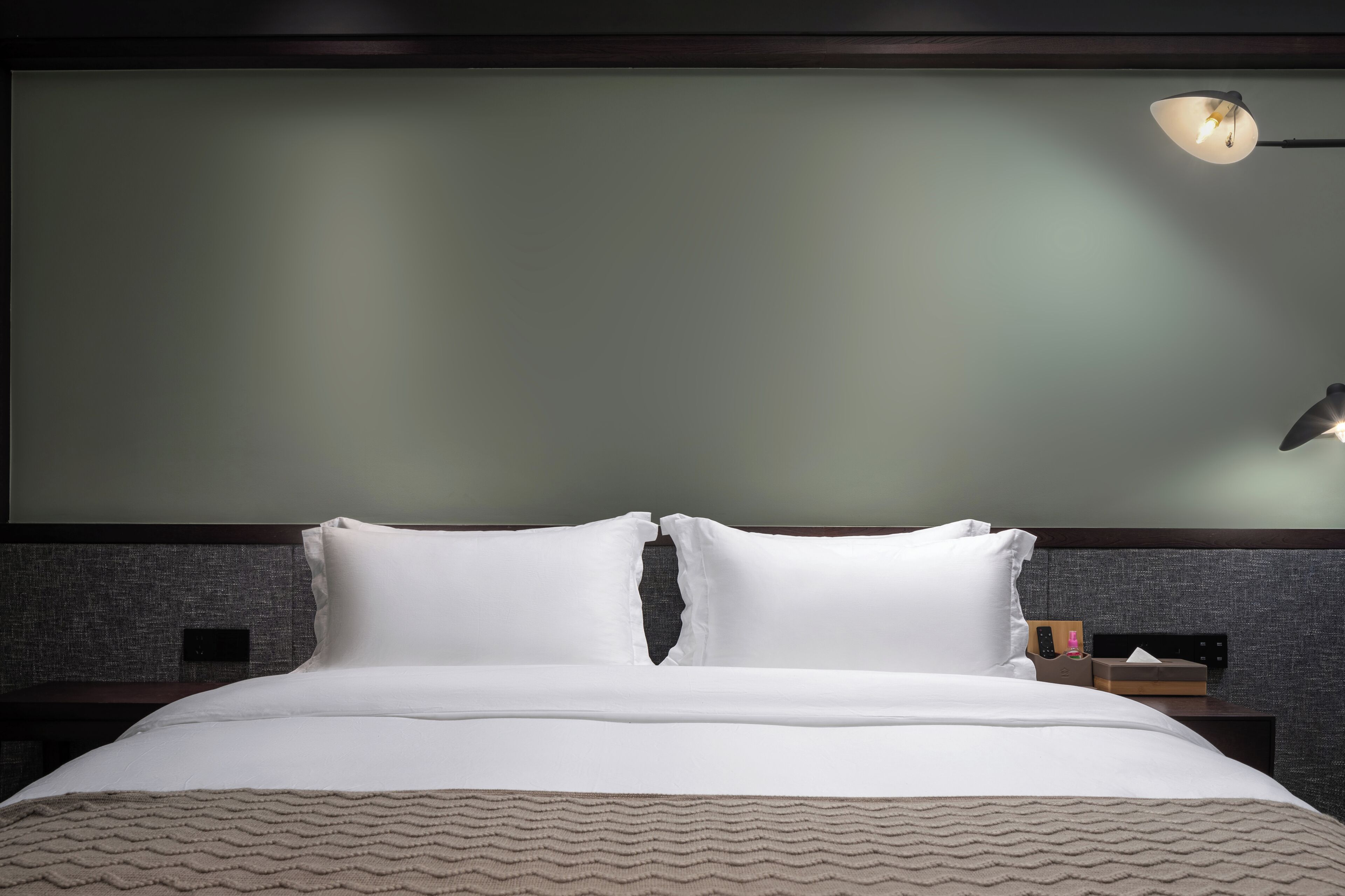 Uma cama bem arrumada com lençóis brancos e manta texturizada, uma mesa de cabeceira com abajur, e um luminário fixado na parede.