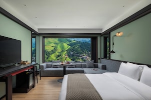 A modern hotel room with a large bed, a sofa, a dining table, and a TV. The room has a large window with a view of green hills and a town.