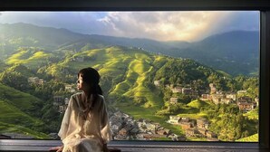 Una persona sentada junto a una ventana con vista a un paisaje escalonado y edificios.