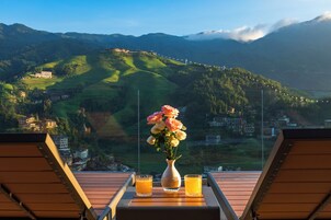 A table with a vase of flowers and two glasses of orange juice, overlooking a mountainous landscape with buildings.