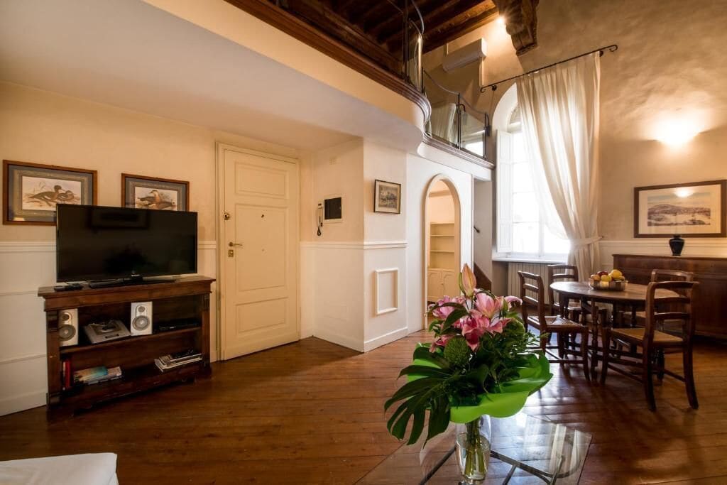 Un soggiorno con televisione, mobile porta TV in legno, zona pranzo con sedie e tavolo, e una scala che conduce a un piano superiore.
