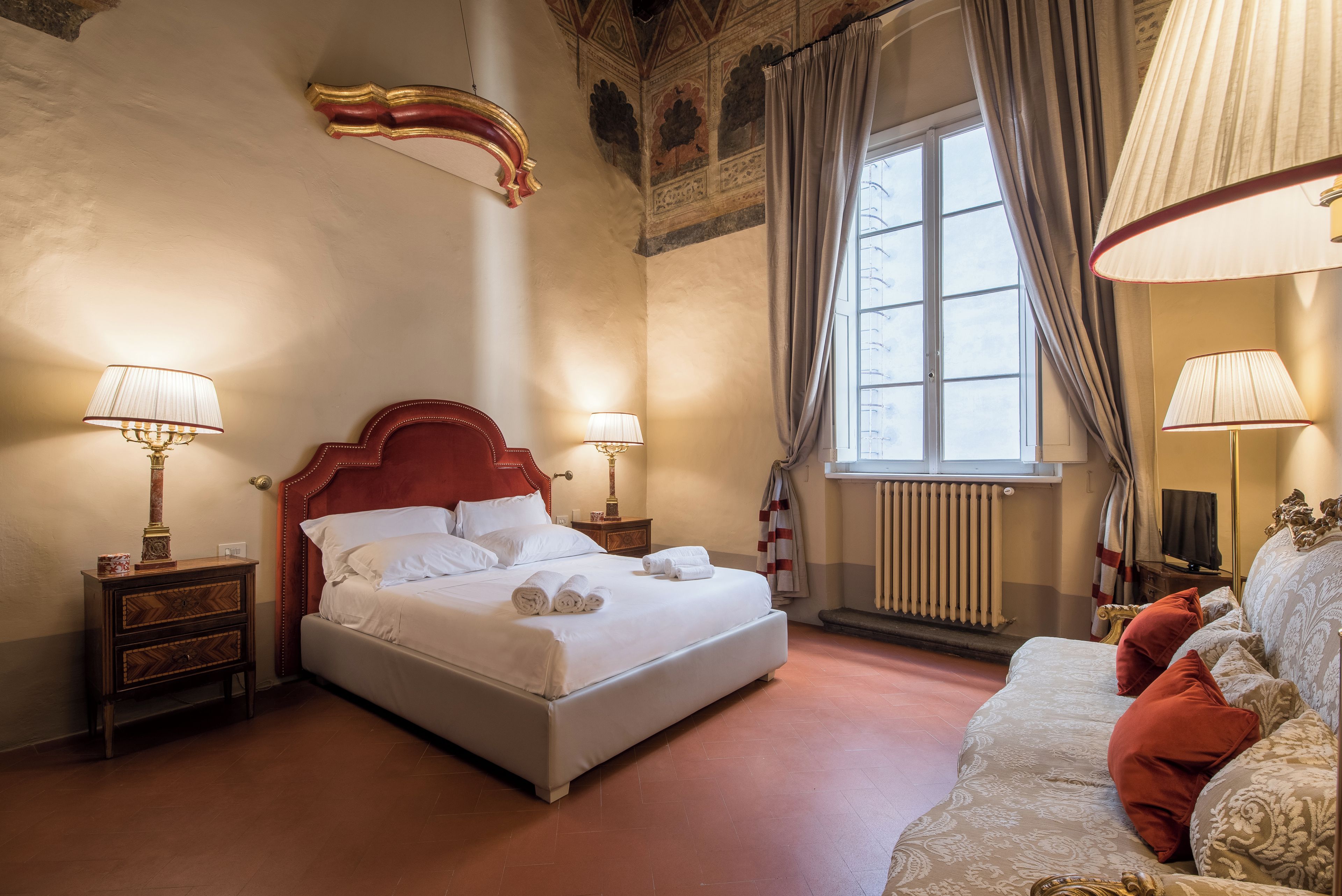 A hotel room with a large bed, two bedside tables with lamps, a window with curtains, a radiator, and a sofa with pillows.