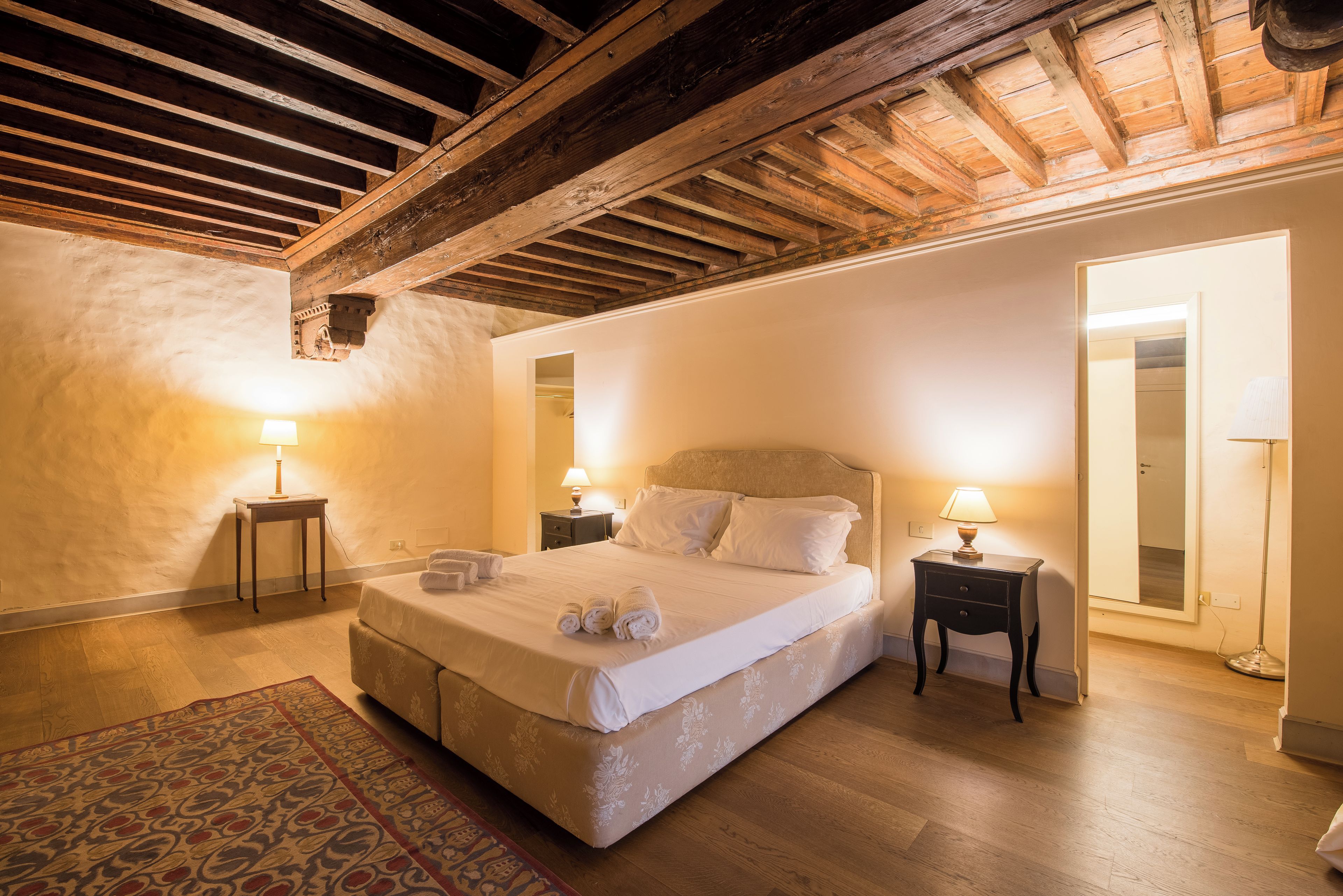 A hotel room with a large bed, two bedside tables with lamps, a patterned rug, and a wooden ceiling.