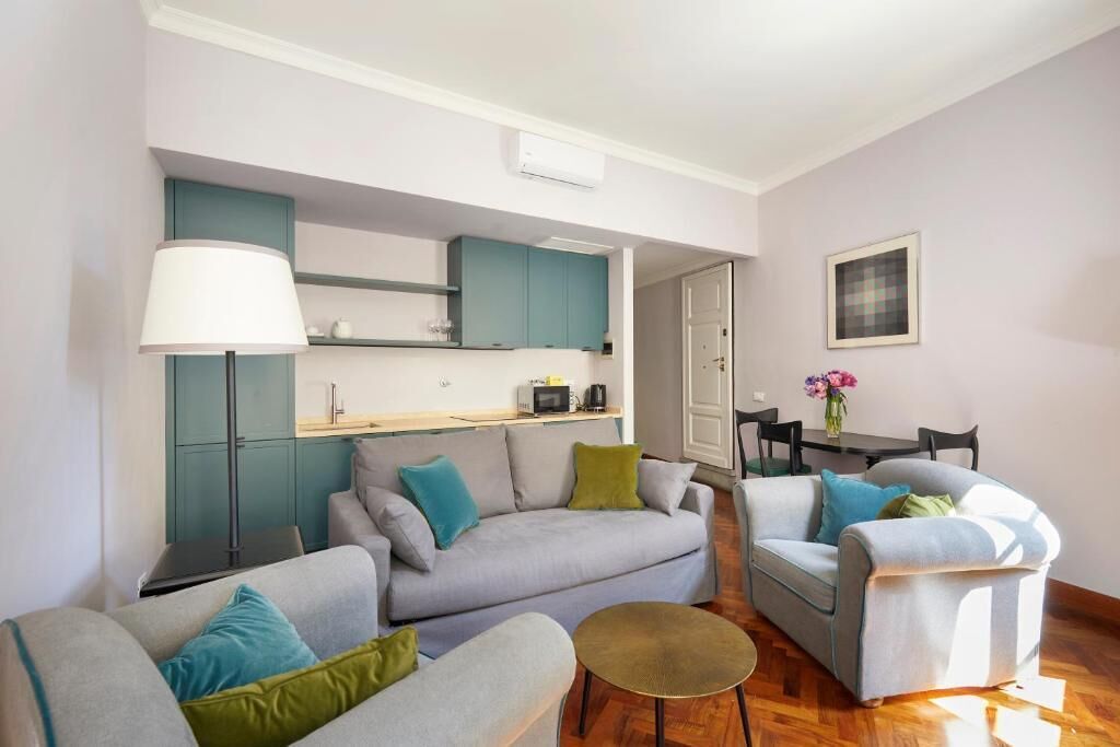 A modern living space with a sofa, armchairs, a small round table, and a kitchenette area.