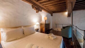 Une pièce avec un plafond en bois, un lit recouvert de draps blancs, une table de chevet avec une lampe et une chaise agrémentée d’un coussin à motifs.