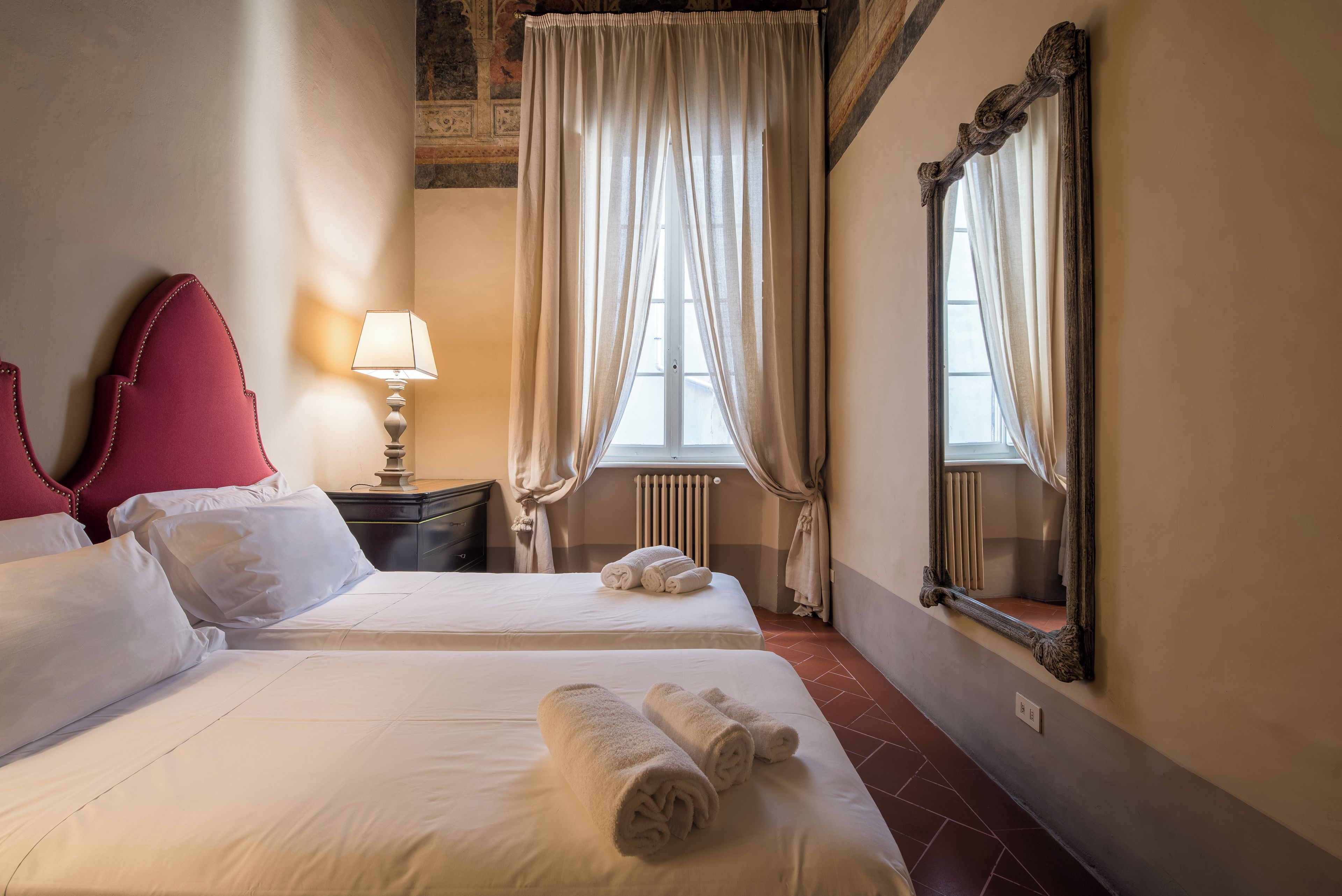 A hotel room with two beds, a nightstand, a lamp, a mirror, and a window with curtains.