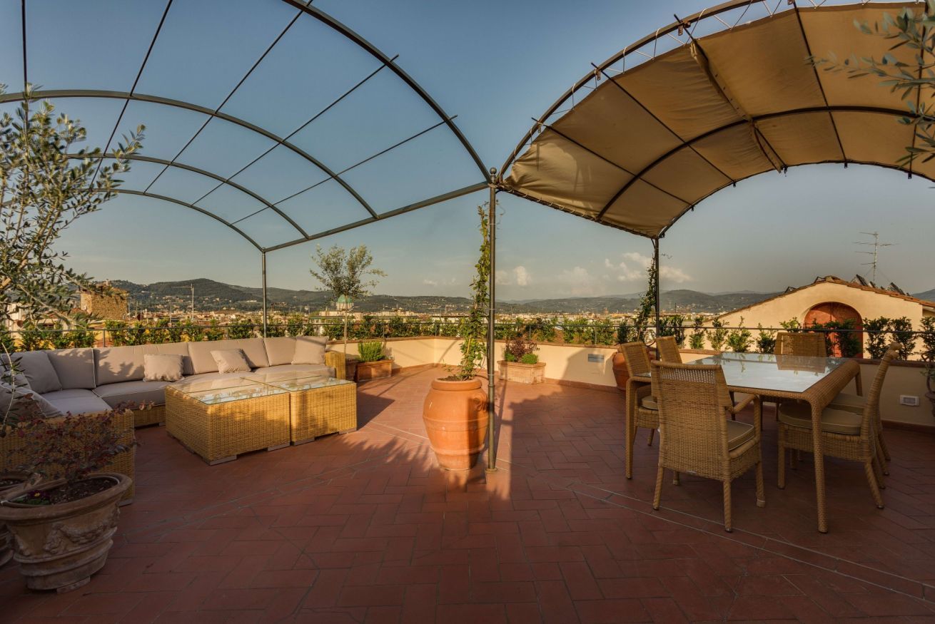 A covered outdoor seating area with a table and chairs, surrounded by potted plants and a view of the landscape.
