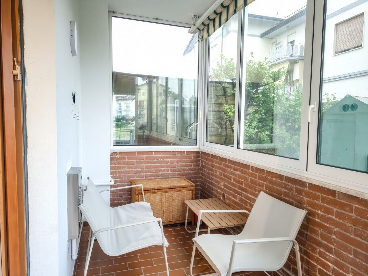 Ein kleiner Balkon mit zwei weißen Stühlen, einem Korbstuhl-Tisch und Blick auf benachbarte Gebäude.
