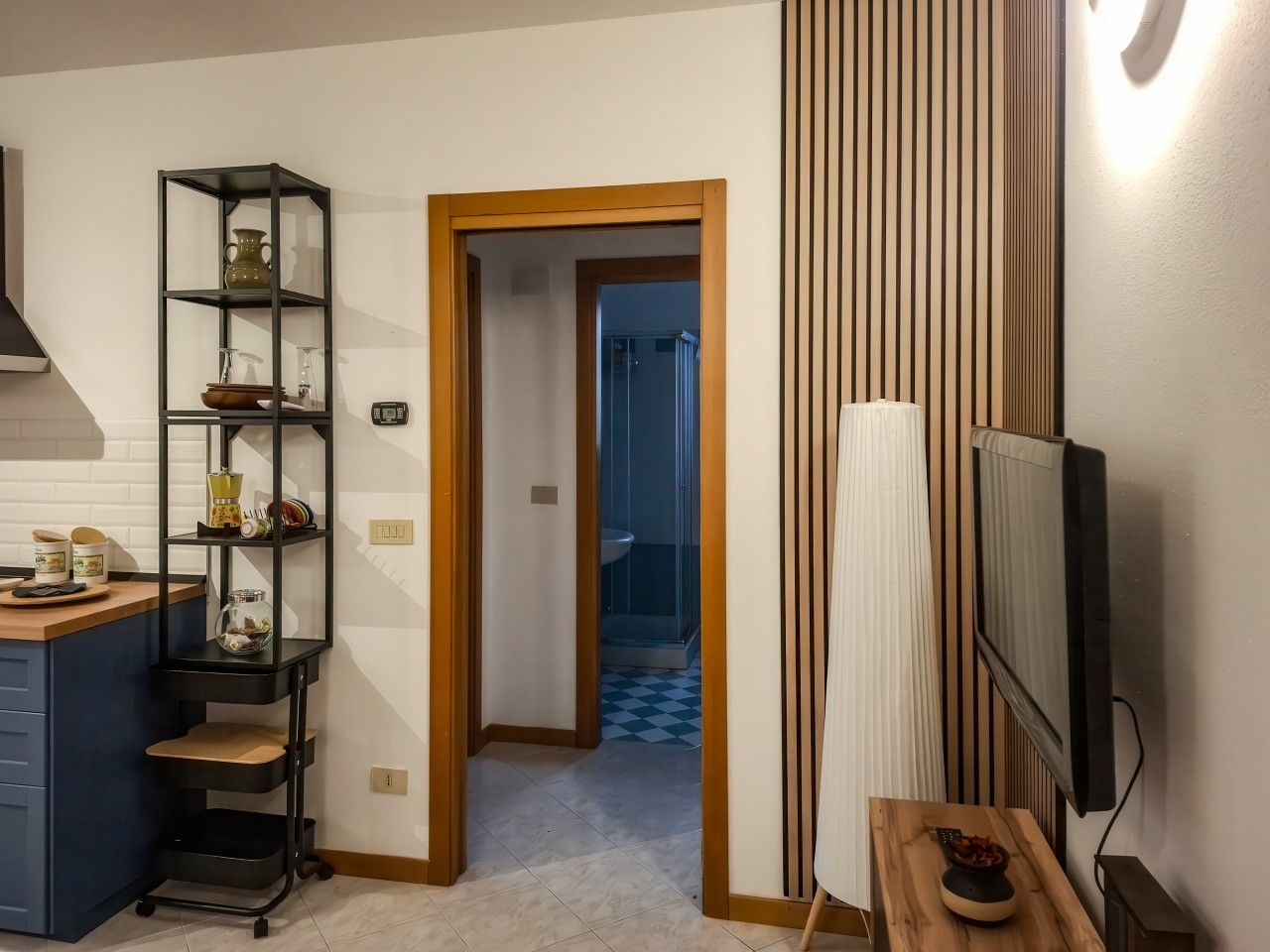 A kitchen with a black metal shelf unit, a wooden door leading to a bathroom, and a television mounted on the wall.