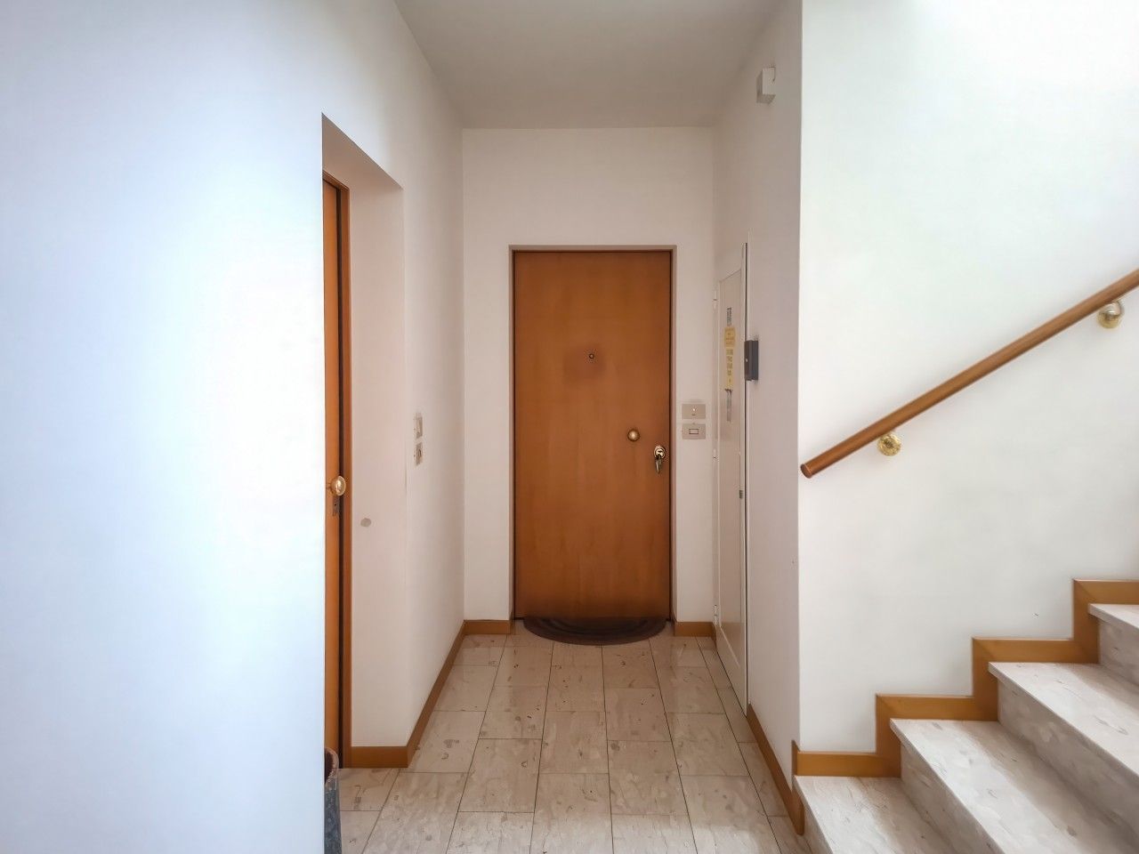 A hallway with a wooden door, a staircase with wooden handrails, and a tiled floor.
