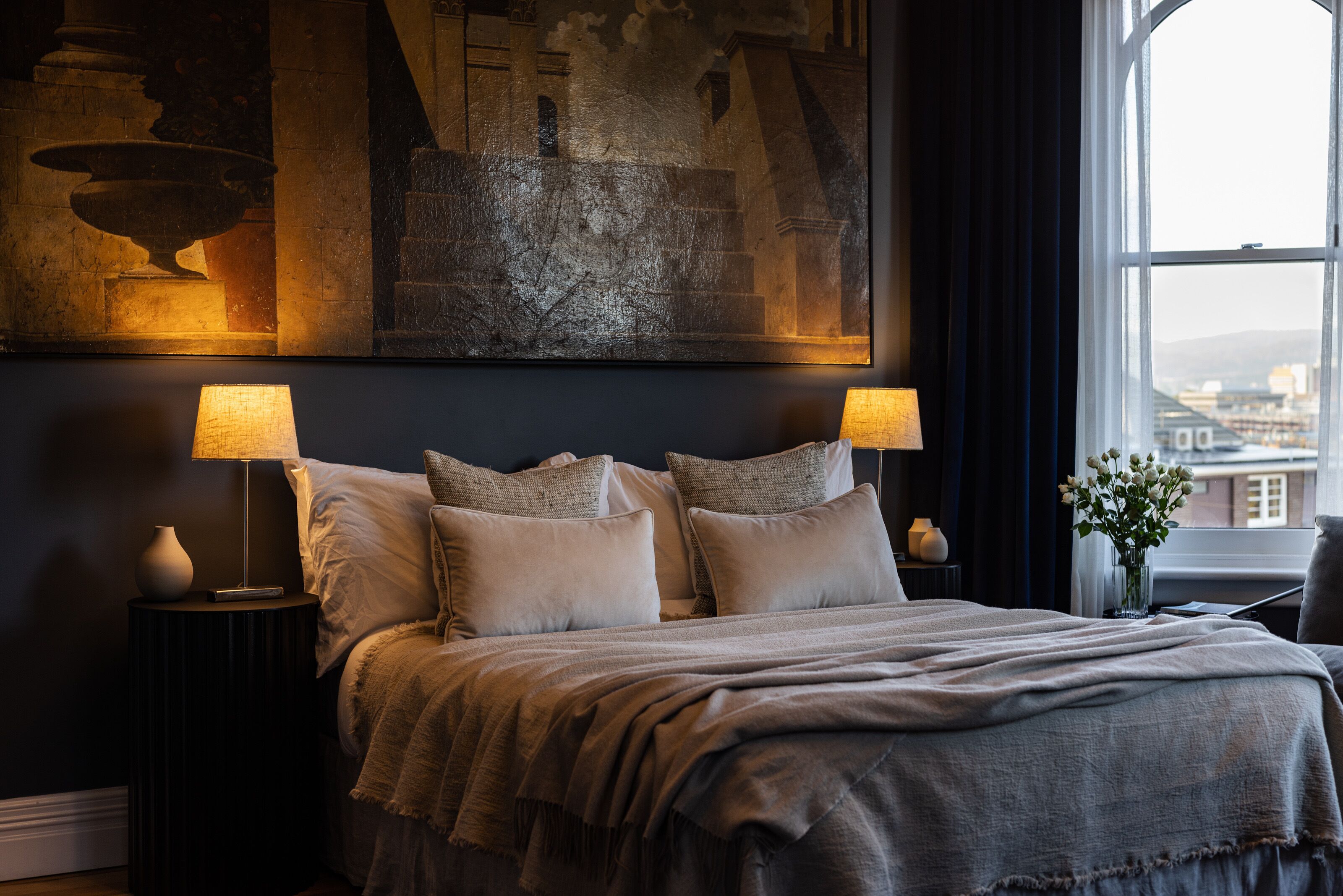 A bedroom with a bed, two bedside lamps, a vase with flowers, and a painting on the wall.