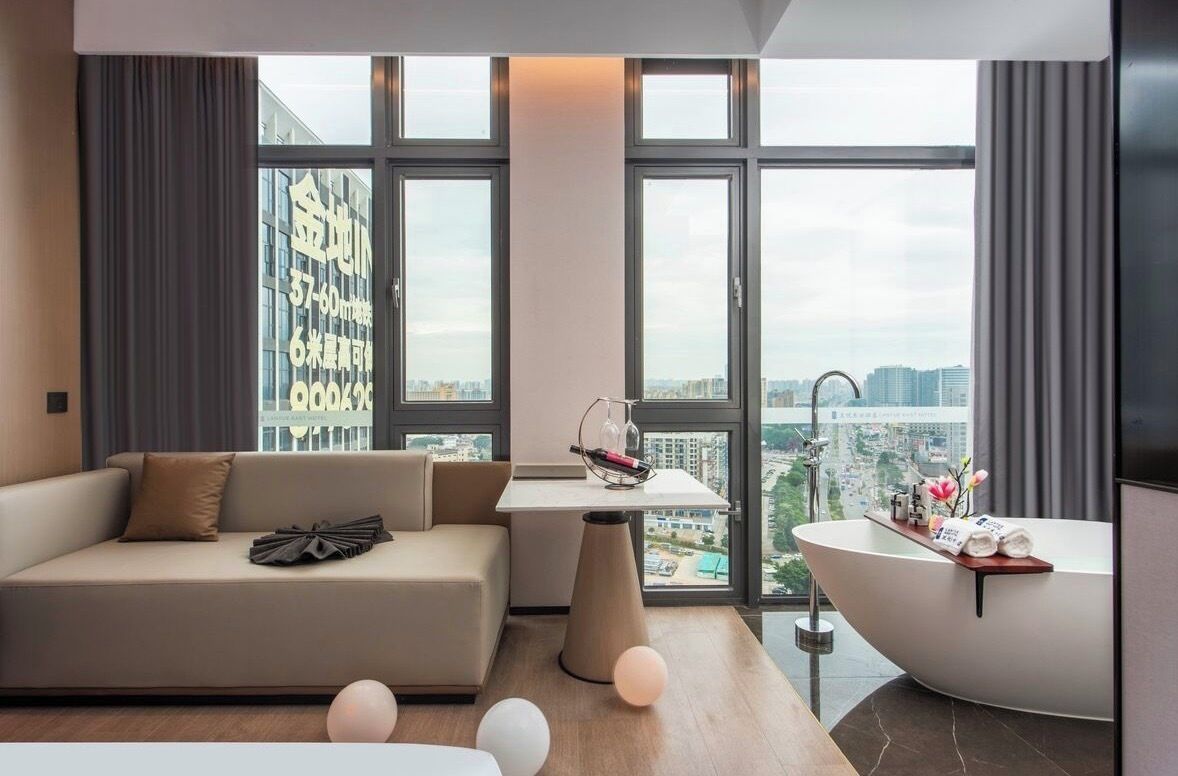 A modern hotel room with a sofa, a small table, a bathtub, and large windows offering a city view.