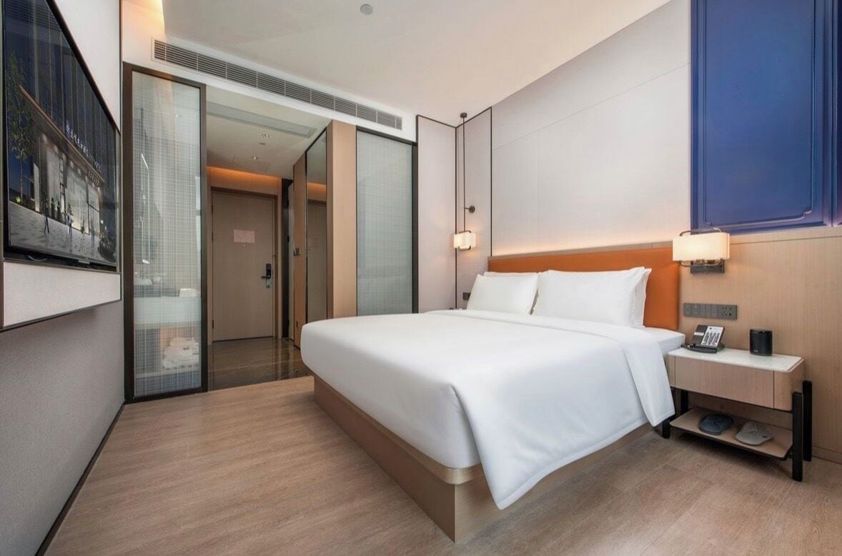 A hotel room with a large bed, a bedside table with a lamp, a wall-mounted TV, and a sliding door leading to a bathroom.