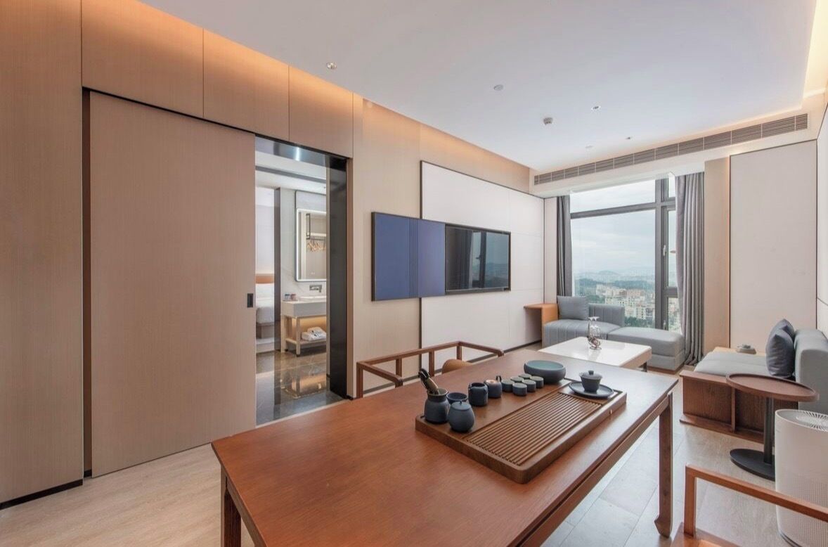 A modern hotel room with a large window, a flat-screen TV, a wooden dining table with tea sets, and a sofa.