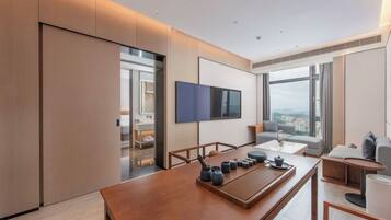 A modern hotel room with a large window, a flat-screen TV, a wooden dining table with tea sets, and a sofa.