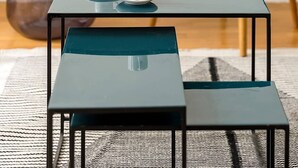 Een kleine, moderne kamer met een donkerblauwe tafel en bank, twee witte kopjes op de tafel, een blauwe fauteuil met een geel kussen, en een hoge, transparante bijzettafel.
