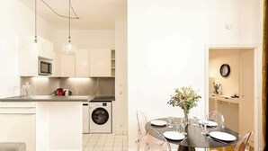 Een moderne keuken met een ingebouwde wasmachine, een eethoek met een ronde tafel en transparante stoelen, en hanglampen.