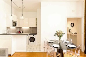 A modern kitchen with a built-in washing machine, a dining area with a round table and clear chairs, and pendant lighting. - Elegant apartment near Place Vendôme - 1 bedroom - long stay - Madeleine I by Habitat Parisien (Paris)