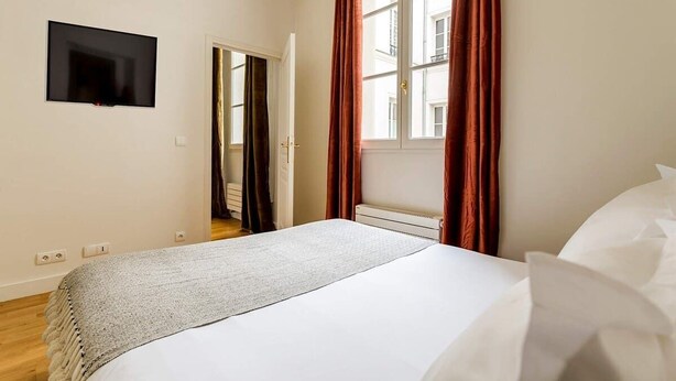 Een slaapkamer met een bed, een aan de muur bevestigde televisie en een raam met gordijnen.