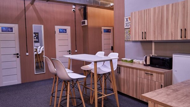 A kitchenette with a microwave, a sink, and wooden cabinets. There is a table with chairs and a door marked with accessibility symbols. - Gagarin Capsule Hotel  (Almaty)