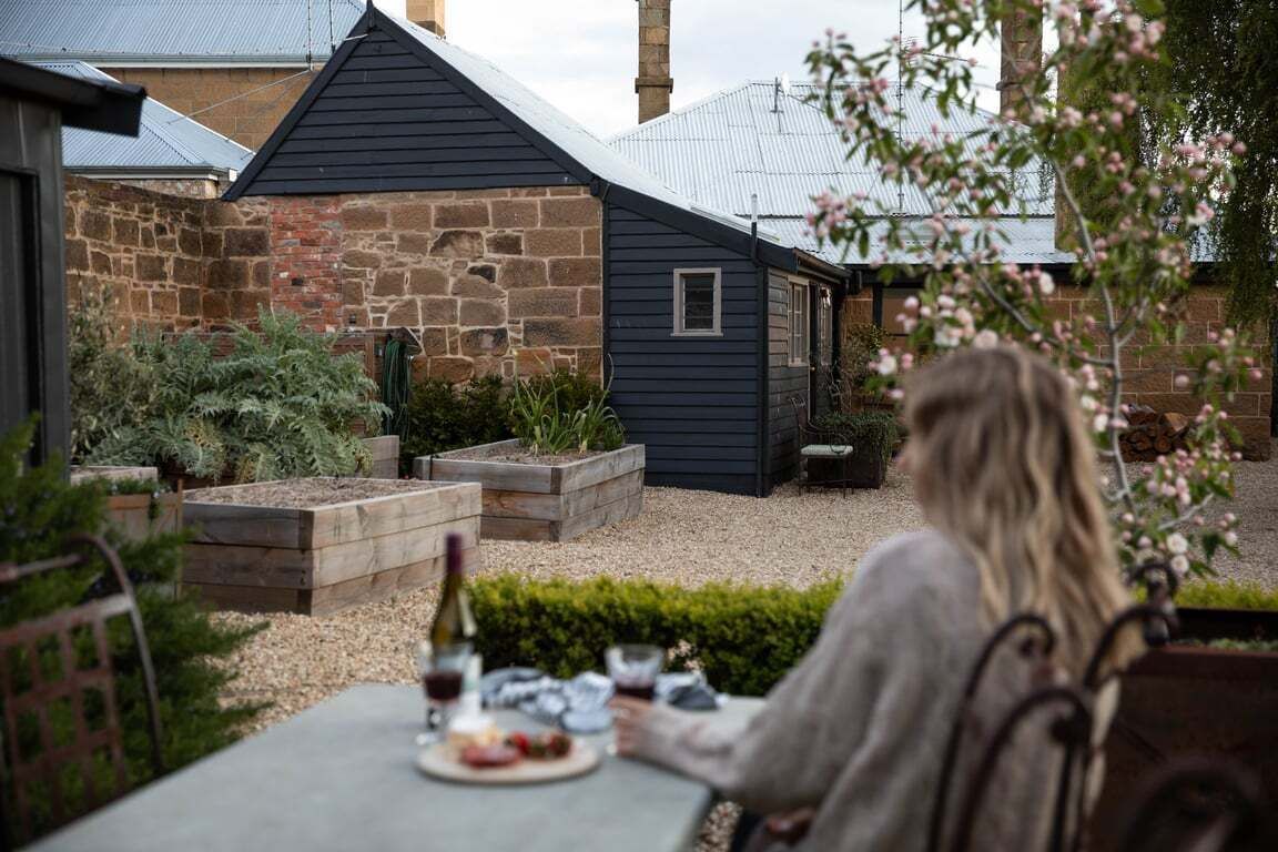 Una persona sentada en una mesa al aire libre con una botella y copas, rodeada de macizos de jardín elevados y un edificio de piedra.