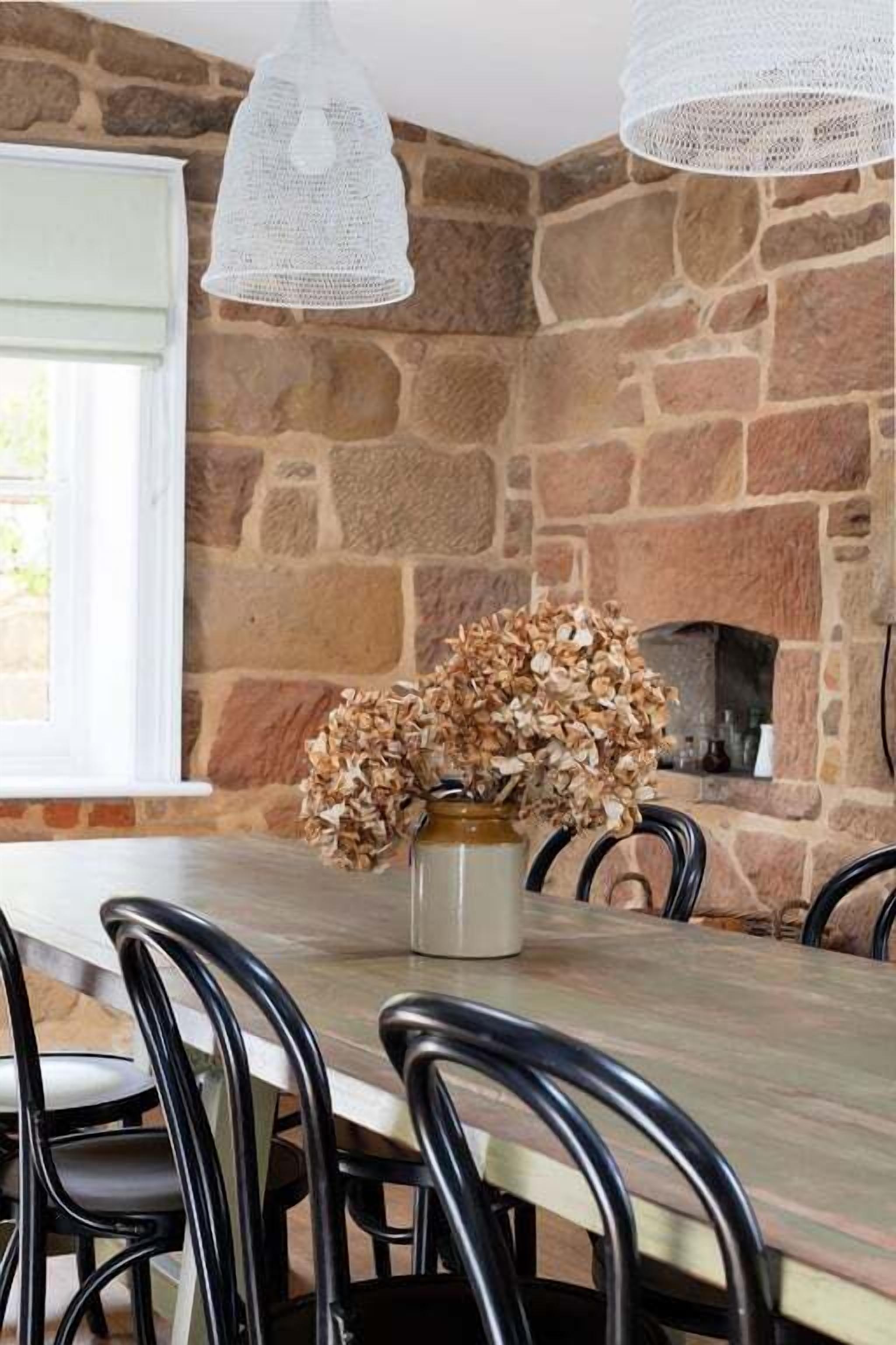 Un coin repas avec un mur en pierre, une table en bois, des chaises noires et un vase contenant des fleurs séchées.