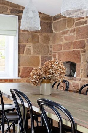 A dining area with a stone wall, a wooden table, black chairs, and a vase with dried flowers.