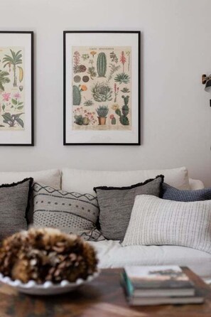 A living room with a sofa, pillows, a coffee table with a book and a bowl of nuts. Two framed botanical prints are hung on the wall.