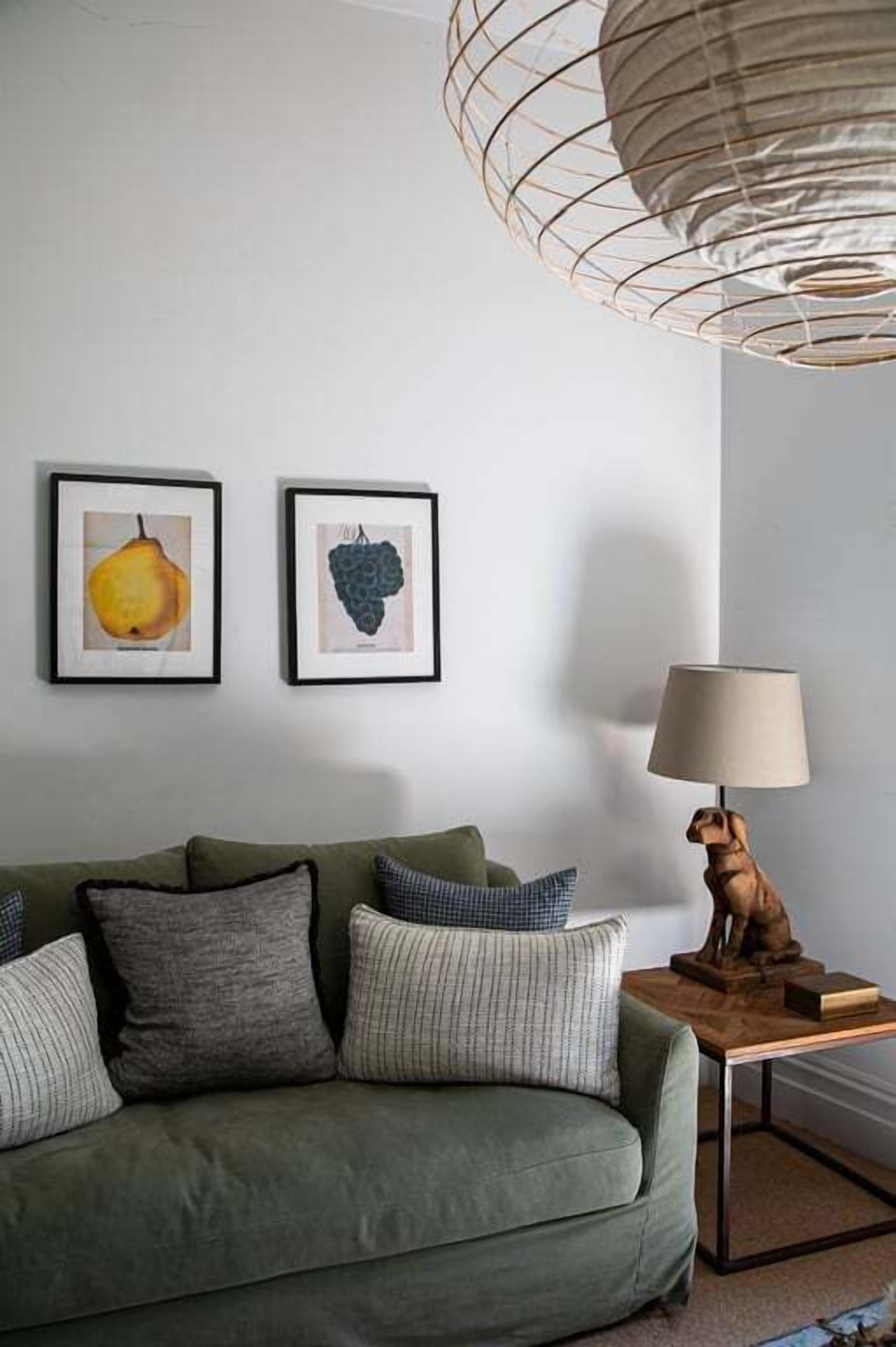 A living room with a green sofa, two framed artworks, a wooden side table with a lamp, and a hanging light fixture.